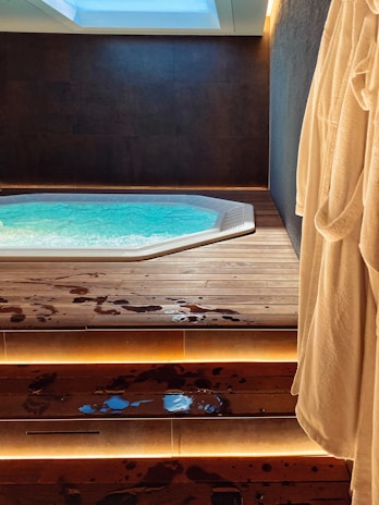A modern indoor hot tub with bubbling water is set in a dark, minimalist room. Wooden steps lead up to the tub, with water droplets scattered across the surface. Soft, indirect lighting highlights the steps, and a plush bathrobe hangs on the right side of the image.