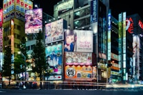 A vibrant cityscape at night featuring numerous illuminated billboards and signs. Many of the signs display colorful anime and manga characters. The scene includes tall buildings densely packed together, lit by bright neon lights, creating a lively and urban atmosphere. Trees line the street, and the street below is busy with moving traffic.