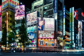 A vibrant cityscape at night featuring numerous illuminated billboards and signs. Many of the signs display colorful anime and manga characters. The scene includes tall buildings densely packed together, lit by bright neon lights, creating a lively and urban atmosphere. Trees line the street, and the street below is busy with moving traffic.