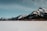 A long freight train with brightly colored containers is moving through a vast snowy landscape with majestic, snow-covered mountains in the background under a clear blue sky.