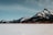 A long freight train with brightly colored containers is moving through a vast snowy landscape with majestic, snow-covered mountains in the background under a clear blue sky.
