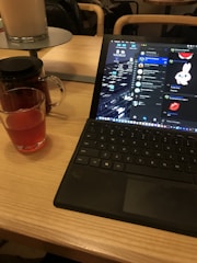 A wooden table holds an open laptop displaying a chat conversation on its screen, alongside a glass cup filled with a red beverage and a teapot nearby. The background shows a dimly lit seating area.