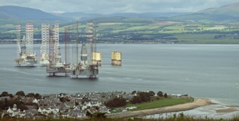 A coastal scene with offshore oil rigs visible in the distance on a calm body of water. In the foreground, there is a town with scattered houses surrounded by green fields. Rolling hills can be seen in the background under a cloudy sky.