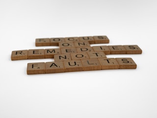 Wooden Scrabble tiles are arranged to form the motivational message: 'FOCUS ON REMEDIES NOT FAULTS'. The tiles are set against a plain, white background.