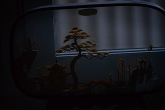 A delicate miniature landscape is encased within a glass frame, depicting a traditional Asian scene with a stylized tree and small architectural structures, including a pagoda and bridges, crafted with intricate details.