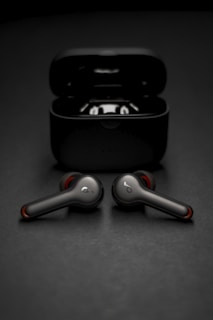 Two wireless earbuds are positioned in front of their open charging case. The earbuds have a sleek, modern design with metallic and red accents. The background is a smooth, dark surface that complements the dark color of the case and earbuds.