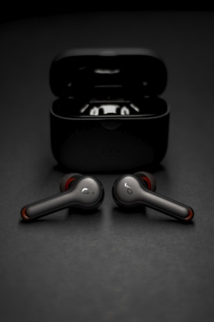 Two wireless earbuds are positioned in front of their open charging case. The earbuds have a sleek, modern design with metallic and red accents. The background is a smooth, dark surface that complements the dark color of the case and earbuds.
