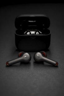 Two wireless earbuds are positioned in front of their open charging case. The earbuds have a sleek, modern design with metallic and red accents. The background is a smooth, dark surface that complements the dark color of the case and earbuds.