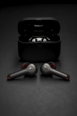 Two wireless earbuds are positioned in front of their open charging case. The earbuds have a sleek, modern design with metallic and red accents. The background is a smooth, dark surface that complements the dark color of the case and earbuds.