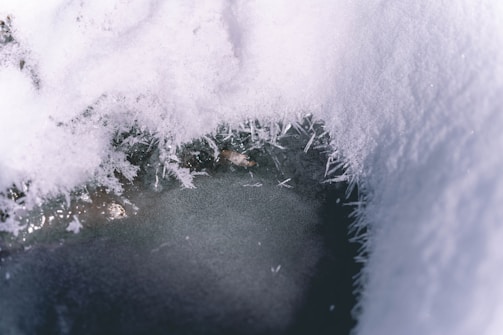 A close-up view of a partially frozen body of water surrounded by snow. Ice crystals and frost form intricate patterns around the edges, creating a textured and delicate appearance.