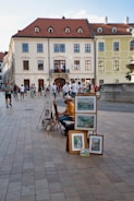 An outdoor scene in a European city square, with a street artist displaying framed paintings and sketches on the pavement. The artist, wearing a yellow shirt, is seated and possibly sketching. The backdrop consists of historic buildings with a red-tiled roof, and there's a group of people casually walking and gathered near a fountain.
