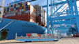 A large container ship is docked at a port with numerous stacked shipping containers on its deck. Blue cranes are positioned along the dock, indicating a busy industrial port environment. The ship and equipment are marked with brand and terminal names.