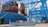 A large container ship is docked at a port with numerous stacked shipping containers on its deck. Blue cranes are positioned along the dock, indicating a busy industrial port environment. The ship and equipment are marked with brand and terminal names.