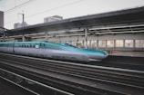 A sleek high-speed train at a modern station platform, with a turquoise and silver color scheme. The platform is covered and features large windows, allowing light to illuminate the interior. Urban buildings can be seen in the background.