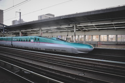 A sleek high-speed train at a modern station platform, with a turquoise and silver color scheme. The platform is covered and features large windows, allowing light to illuminate the interior. Urban buildings can be seen in the background.