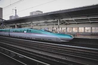 A sleek high-speed train at a modern station platform, with a turquoise and silver color scheme. The platform is covered and features large windows, allowing light to illuminate the interior. Urban buildings can be seen in the background.