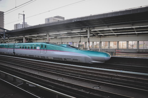 A sleek high-speed train at a modern station platform, with a turquoise and silver color scheme. The platform is covered and features large windows, allowing light to illuminate the interior. Urban buildings can be seen in the background.