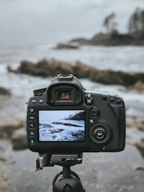 A digital camera mounted on a tripod is focused on a scenic seascape, capturing waves crashing against rocks. The camera's screen displays an image of the ocean, showing a similar scene to the one in the background.