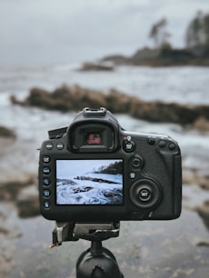 A digital camera mounted on a tripod is focused on a scenic seascape, capturing waves crashing against rocks. The camera's screen displays an image of the ocean, showing a similar scene to the one in the background.