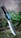 A knife with a textured handle and a metallic blade is embedded in the ground. The handle features dark brown and blue hues with small visible rivets. Surrounding the knife are blurry green plants and grass, giving a natural and rustic setting.
