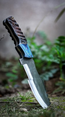 A knife with a textured handle and a metallic blade is embedded in the ground. The handle features dark brown and blue hues with small visible rivets. Surrounding the knife are blurry green plants and grass, giving a natural and rustic setting.