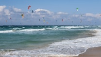 A sandy beach with gentle waves rolling onto the shore is depicted, accompanied by numerous colorful kitesurfing kites scattered across the sky. The sky is partly cloudy with patches of blue, and the ocean appears to be a rich blue-green color.
