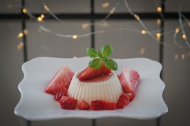 A creamy panna cotta dessert is elegantly presented on a square white plate, garnished with fresh strawberry slices and topped with a small sprig of green mint. In the background, there are soft, warm decorative string lights creating a cozy and inviting atmosphere.