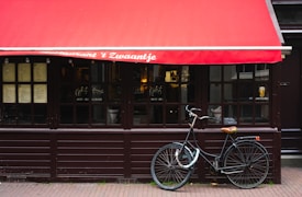 A quaint restaurant facade features a red awning with white text, large wooden-framed windows displaying menus, and a black bicycle parked in front on the brick-paved sidewalk.