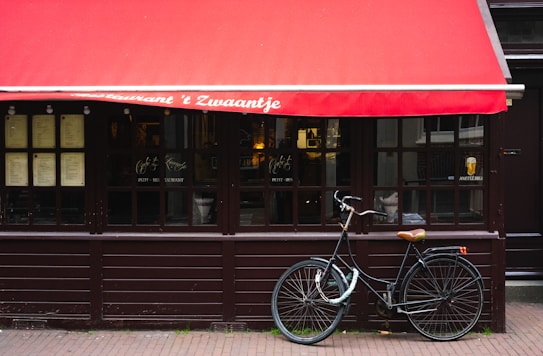 A quaint restaurant facade features a red awning with white text, large wooden-framed windows displaying menus, and a black bicycle parked in front on the brick-paved sidewalk.