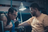 Two men are in a dimly lit room, with one giving a tattoo to the other. The tattoo artist is focused on his work, holding a tattoo machine and wearing gloves. The client sits patiently, extending his arm for the tattoo. A lamp hangs above, illuminating the work area. Various tattoo equipment and supplies are visible in the background.
