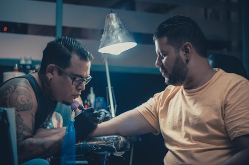 Two men are in a dimly lit room, with one giving a tattoo to the other. The tattoo artist is focused on his work, holding a tattoo machine and wearing gloves. The client sits patiently, extending his arm for the tattoo. A lamp hangs above, illuminating the work area. Various tattoo equipment and supplies are visible in the background.