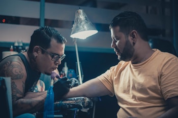 Two men are in a dimly lit room, with one giving a tattoo to the other. The tattoo artist is focused on his work, holding a tattoo machine and wearing gloves. The client sits patiently, extending his arm for the tattoo. A lamp hangs above, illuminating the work area. Various tattoo equipment and supplies are visible in the background.