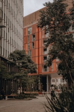 A modern urban setting featuring two tall buildings with distinct architectural designs. One building has a sleek, metallic facade, while the other showcases a warm, orange-toned exterior. Lush green trees line the foreground, offering a natural contrast to the urban environment. A person is walking along the pathway, surrounded by planters and a well-maintained courtyard area.