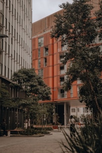 A modern urban setting featuring two tall buildings with distinct architectural designs. One building has a sleek, metallic facade, while the other showcases a warm, orange-toned exterior. Lush green trees line the foreground, offering a natural contrast to the urban environment. A person is walking along the pathway, surrounded by planters and a well-maintained courtyard area.