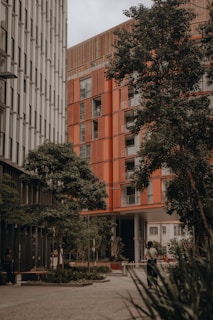A modern urban setting featuring two tall buildings with distinct architectural designs. One building has a sleek, metallic facade, while the other showcases a warm, orange-toned exterior. Lush green trees line the foreground, offering a natural contrast to the urban environment. A person is walking along the pathway, surrounded by planters and a well-maintained courtyard area.