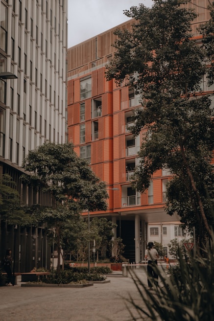 A modern urban setting featuring two tall buildings with distinct architectural designs. One building has a sleek, metallic facade, while the other showcases a warm, orange-toned exterior. Lush green trees line the foreground, offering a natural contrast to the urban environment. A person is walking along the pathway, surrounded by planters and a well-maintained courtyard area.