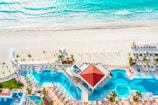 An aerial view of a luxurious resort featuring two large swimming pools with clear blue water, surrounded by numerous lounge chairs and umbrellas. The resort is situated right next to an expansive sandy beach with turquoise waves gently crashing onto the shore. Palm trees are scattered around, enhancing the tropical vibe.