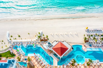 An aerial view of a luxurious resort featuring two large swimming pools with clear blue water, surrounded by numerous lounge chairs and umbrellas. The resort is situated right next to an expansive sandy beach with turquoise waves gently crashing onto the shore. Palm trees are scattered around, enhancing the tropical vibe.