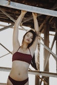 A person in a maroon bikini stands underneath a metal structure, with arms raised to hold onto a beam. The sunlight creates a warm backlit effect, enhancing the overall mood.