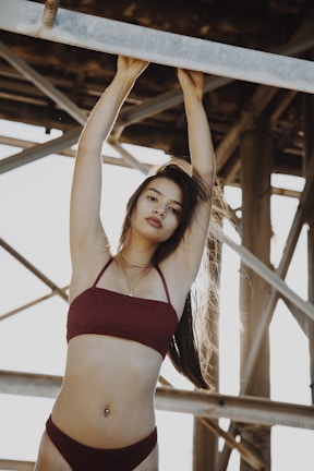 A person in a maroon bikini stands underneath a metal structure, with arms raised to hold onto a beam. The sunlight creates a warm backlit effect, enhancing the overall mood.