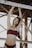 A person in a maroon bikini stands underneath a metal structure, with arms raised to hold onto a beam. The sunlight creates a warm backlit effect, enhancing the overall mood.