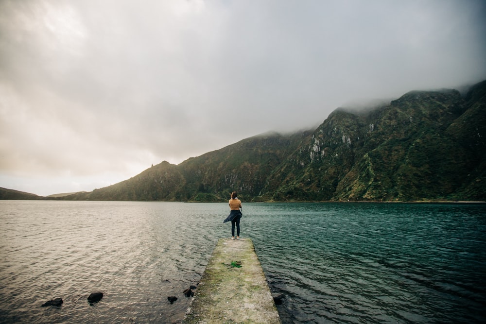 woman in blue jacket and black pants standing on dock near body of water during daytime