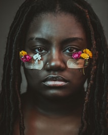 A person with long hair adorned with small, colorful flowers is gazing directly at the viewer. Patches with the words 'TAKE CARE' and 'OF YOURSELF' are placed under their eyes, emphasizing a message of self-care.