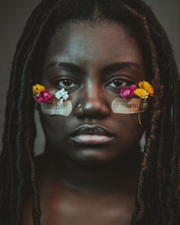 A person with long hair adorned with small, colorful flowers is gazing directly at the viewer. Patches with the words 'TAKE CARE' and 'OF YOURSELF' are placed under their eyes, emphasizing a message of self-care.