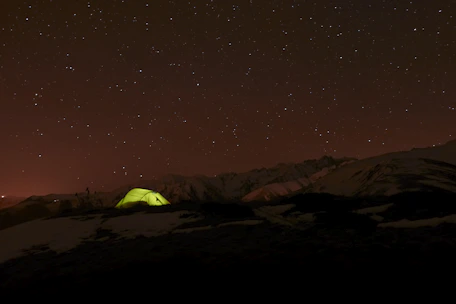 A local guide sharing ancient Himalayan stories around a campfire at dusk, with tents and mountains silhouetted behind.