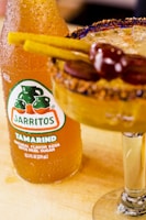 A glass bottle of Jarritos Tamarind soda stands beside a cocktail glass. The cocktail is garnished with a spicy rim, tamarind sticks, and cherries. The beverage is golden and refreshing, with condensation on the bottle indicating a chilled drink.