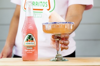 A bottle of Jarritos guava soda is placed on a wooden surface. A person is holding a margarita glass filled with a pink beverage, topped with a slush and a blue sugar rim. The person is wearing a shirt with a partially visible logo that matches the bottle design.