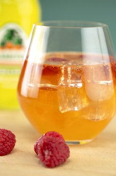 A glass filled with a refreshing orange-colored beverage, garnished with ice cubes, is placed on a tabletop. Nearby, a couple of fresh raspberries add a touch of vibrant red. In the background, there is a blurred yellow bottle, suggesting a drink mix or juice.