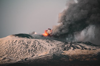 A volcanic eruption with fiery lava and dense smoke billowing into the sky over a snow-capped mountain landscape. The contrast between the white snow and the dark volcanic ash creates a dramatic scene.
