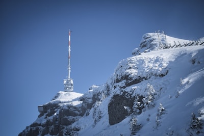 A snowy mountaintop with a large communication tower rising above it. The sky is a clear blue, and the mountain is covered in snow with exposed rocky surfaces. Snow fences are visible along the slope.